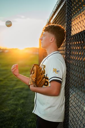 Baseball player in white uniform practices catching drills against chain link fence during golden sunset.