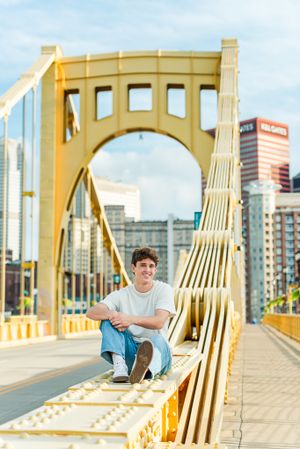 Someone sits casually on yellow Roberto Clemente Bridge railing with downtown Pittsburgh buildings in background.