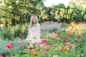A woman in a white dress stands in a summer garden filled with vibrant pink and orange poppies at sunset.