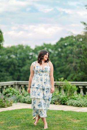 A person in a floral blue and white summer dress walks gracefully through a lush garden setting on a sunny day.