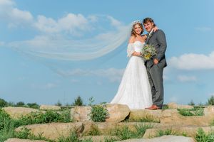 A bride and groom share a romantic moment standing on rocks against a blue sky and green landscape background.