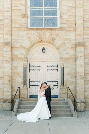 A couple shares a romantic moment on stone steps in front of an elegant arched church doorway with limestone walls.
