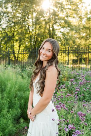 A person in a white dress poses in a garden with purple wildflowers and green foliage during golden hour.