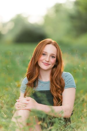 Woman with long red hair wearing a striped shirt and overalls sits in a sunny meadow surrounded by wildflowers.