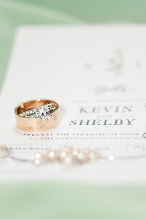Wedding invitation with names Kevin and Shelby alongside diamond engagement ring and delicate pearl jewelry.