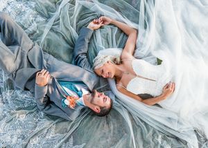 A bride and groom lay on the ground laughing together surrounded by flowing blue and white dress fabric.
