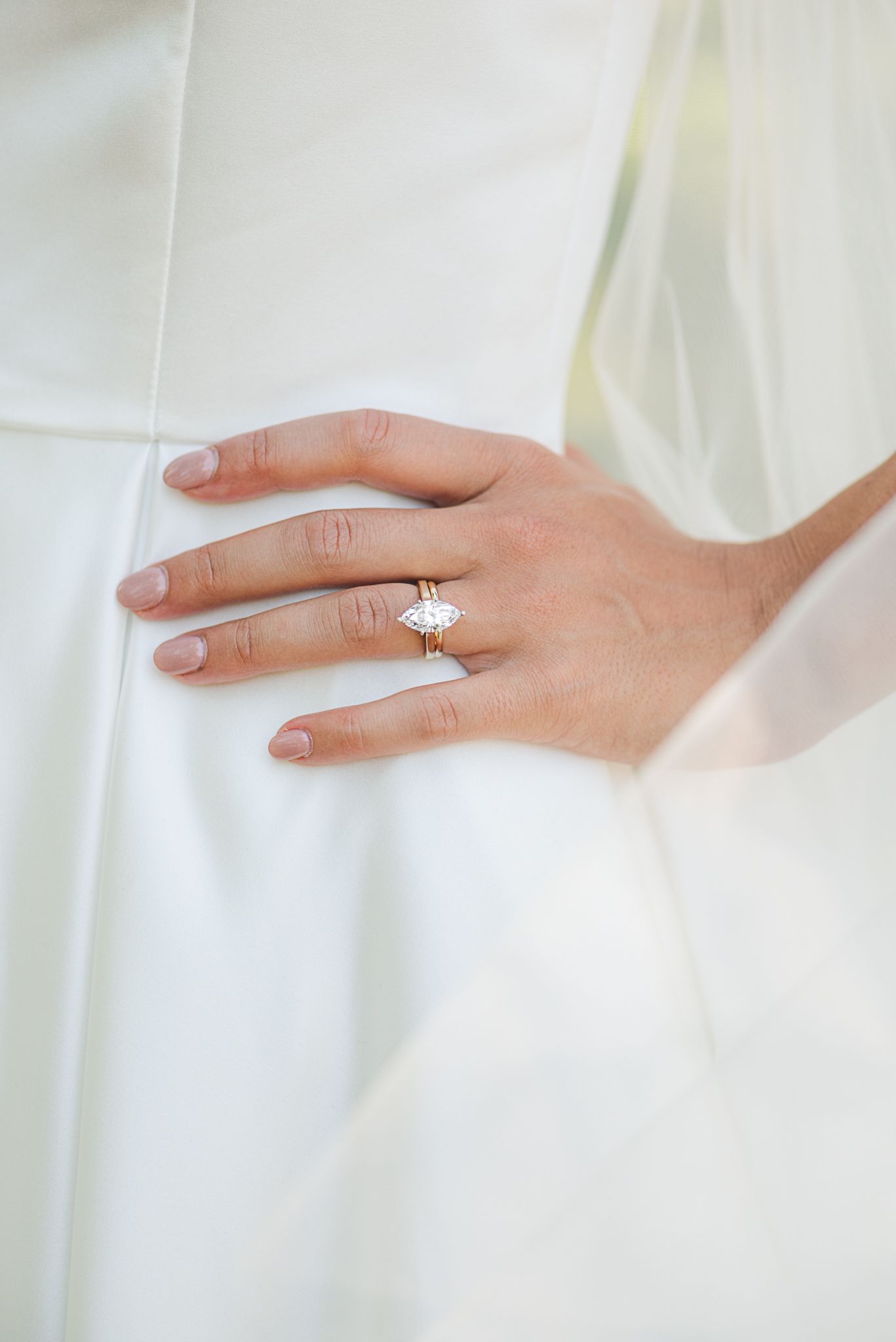Close-up of a delicate diamond engagement ring on a bride's hand resting against white fabric.