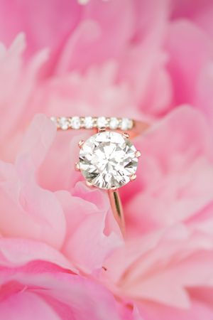 A sparkling diamond engagement ring sits nestled among soft pink peony petals in a romantic close-up shot.