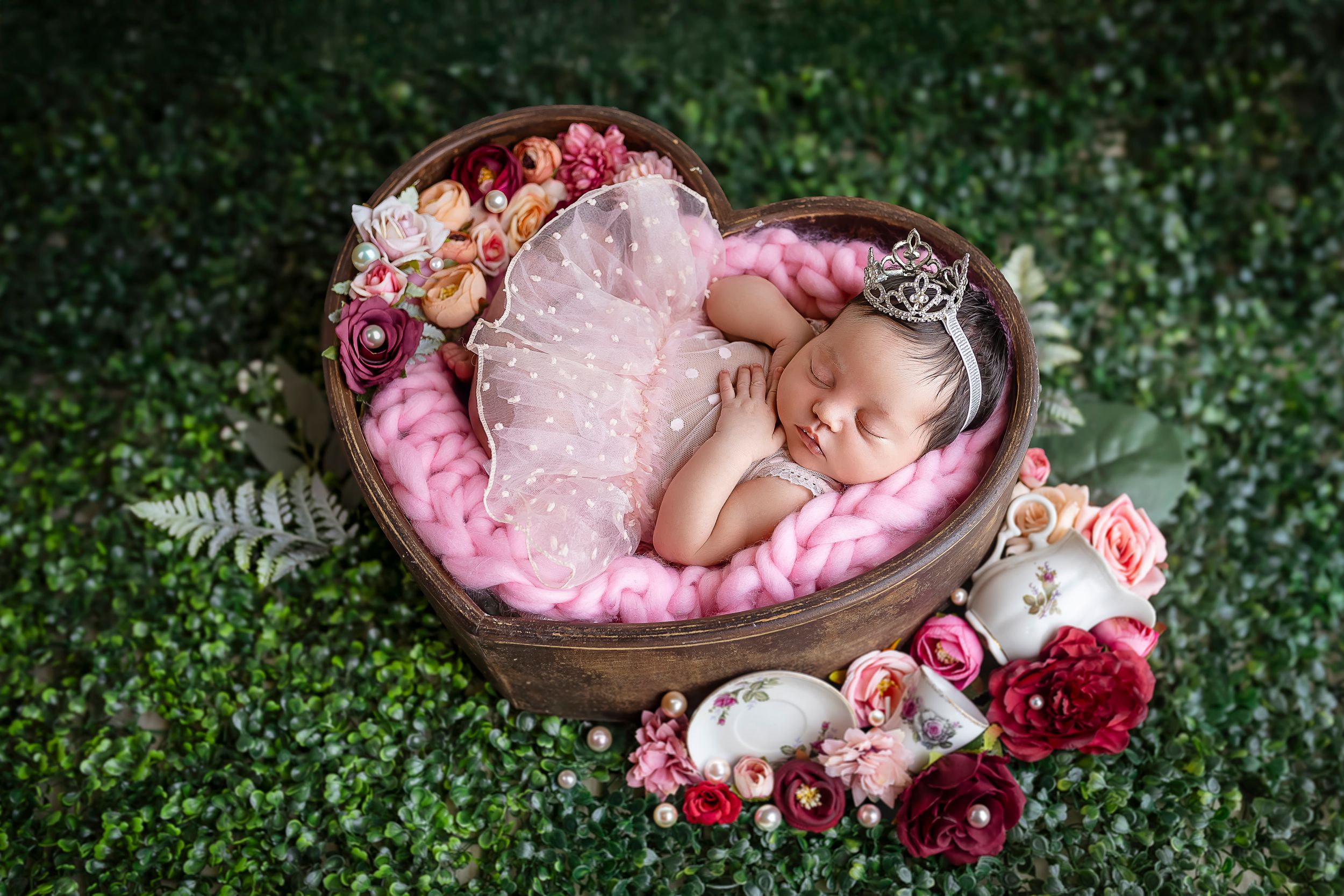 bebe posada dentro de un corazon con un trajecito rosa estilo tutu hay flores y grama verde en el escenario