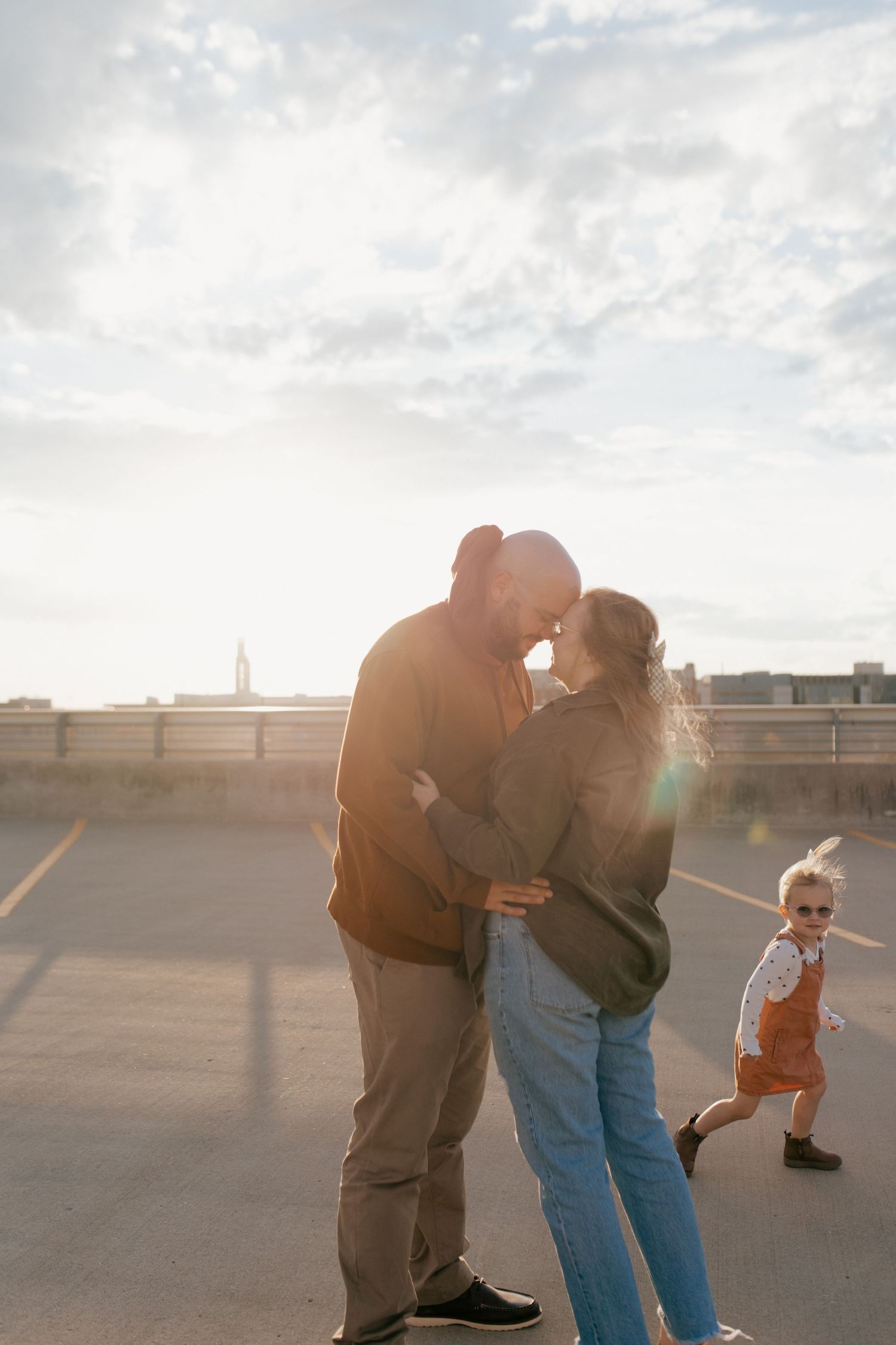 A couple embraces and shares a tender moment at sunset on a rooftop with dramatic lighting and cloudy skies.