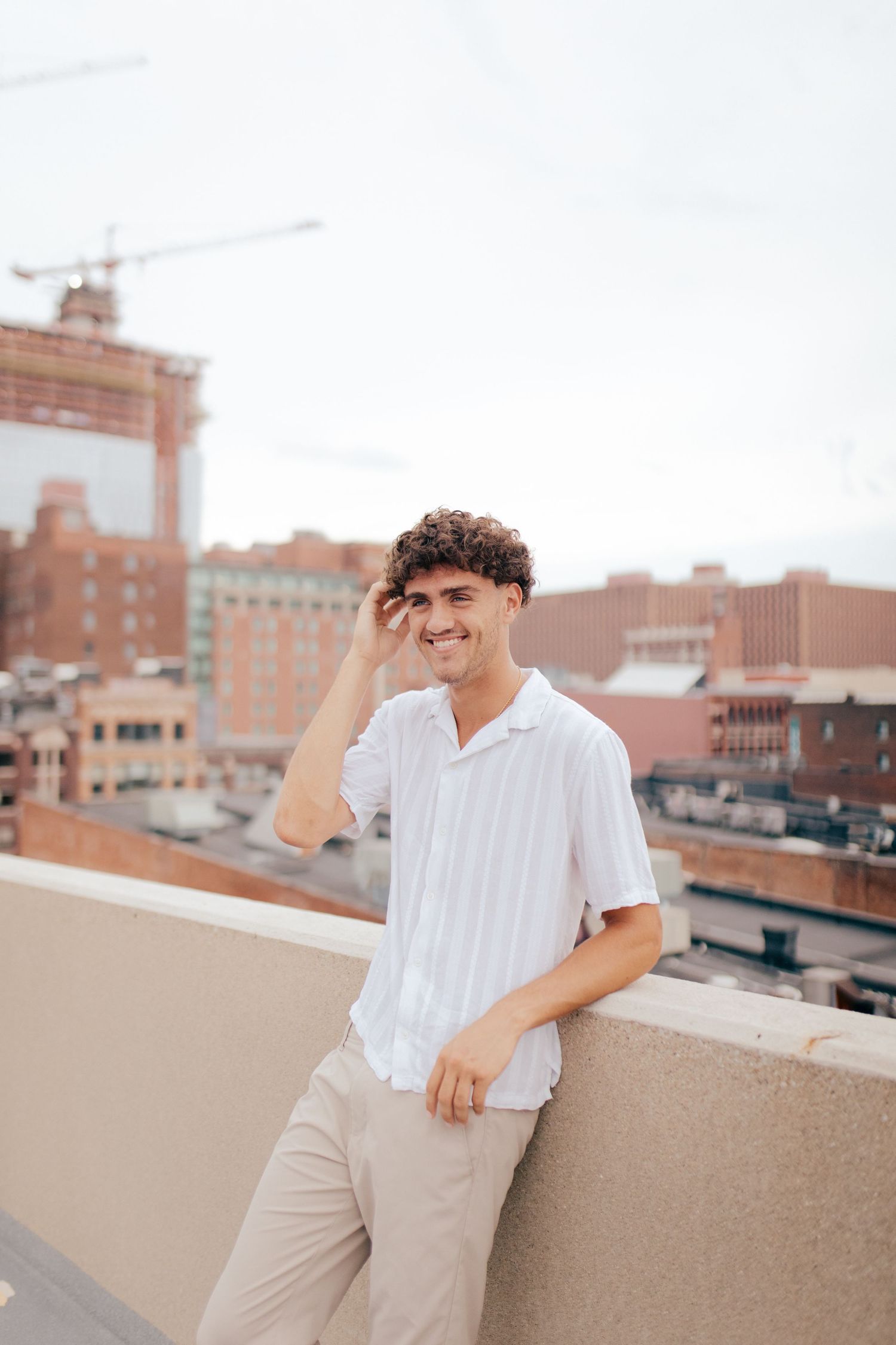 Young person in white button-down shirt and khaki pants smiling on urban rooftop with city buildings in background.