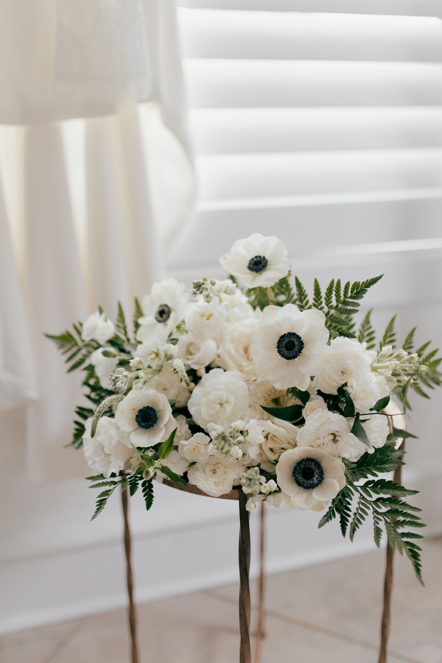 Three elegant white floral centerpieces with anemones, roses and ferns arranged on pedestals against a bright window.