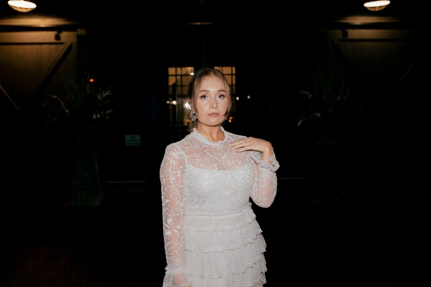 A bride in a white lace gown poses in a dimly lit setting with dramatic lighting.