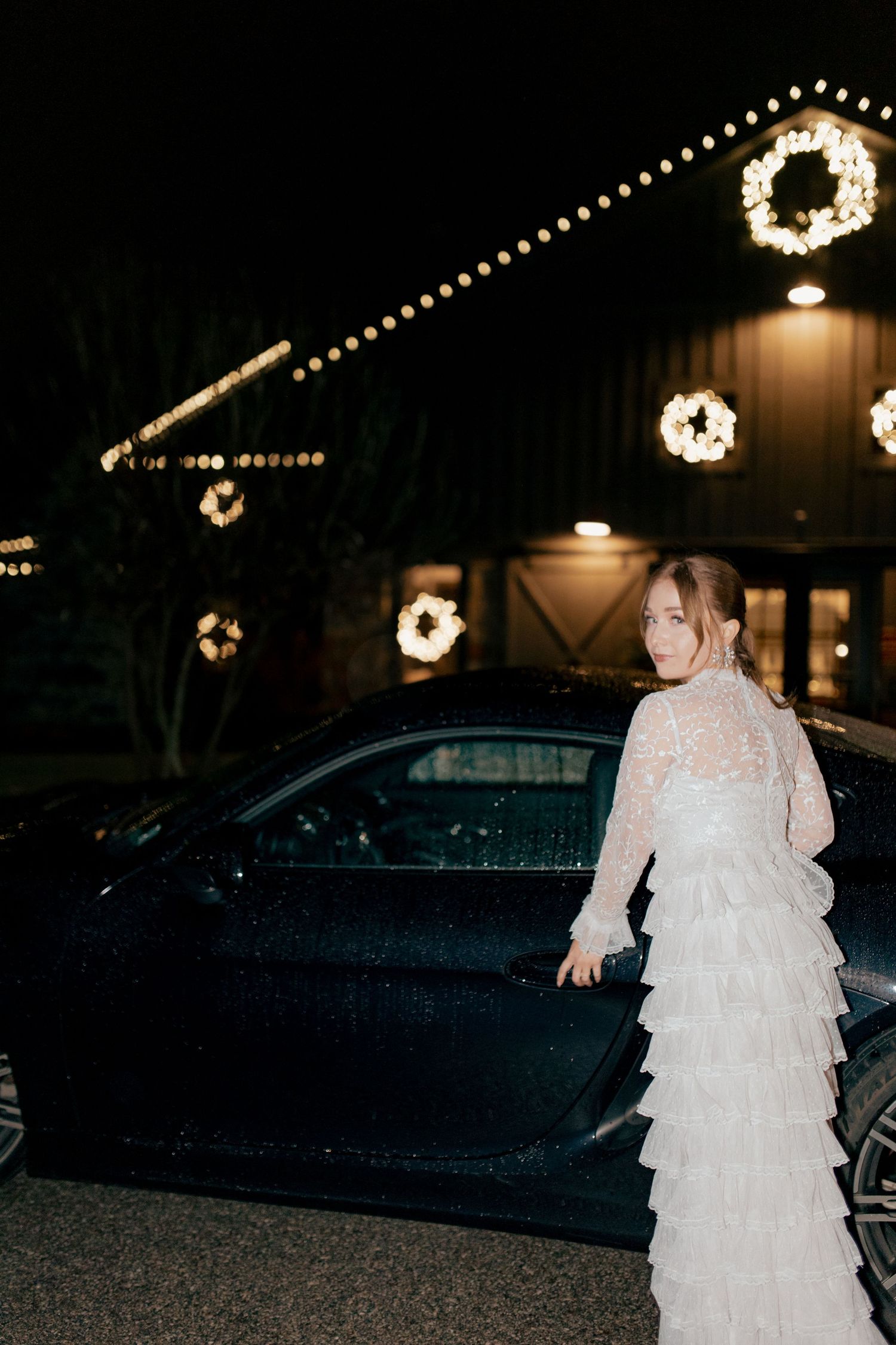 A white lace dress glows under illuminated Christmas wreaths and string lights at a nighttime outdoor venue.