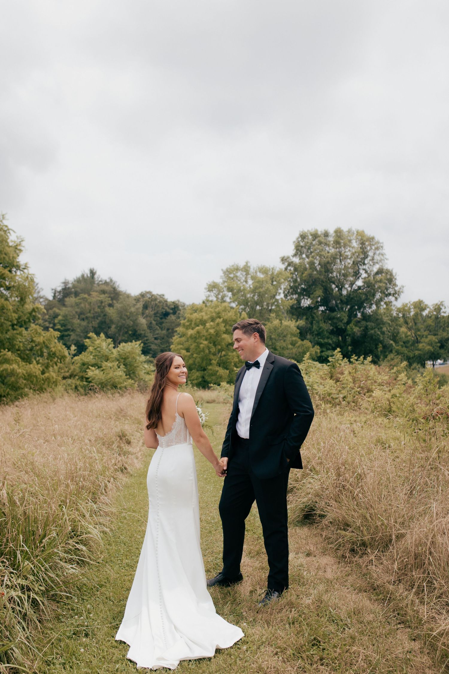 A couple in wedding attire shares a joyful moment together in a rustic field with tall grass and trees in the background.