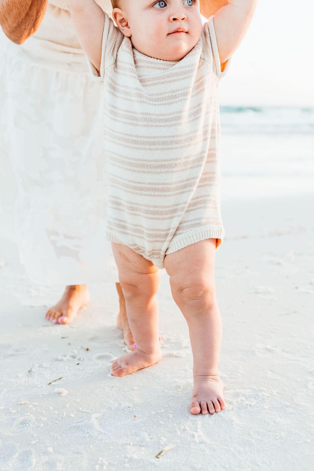 Small child in striped romper taking first steps on sandy beach while supported by parent hands.