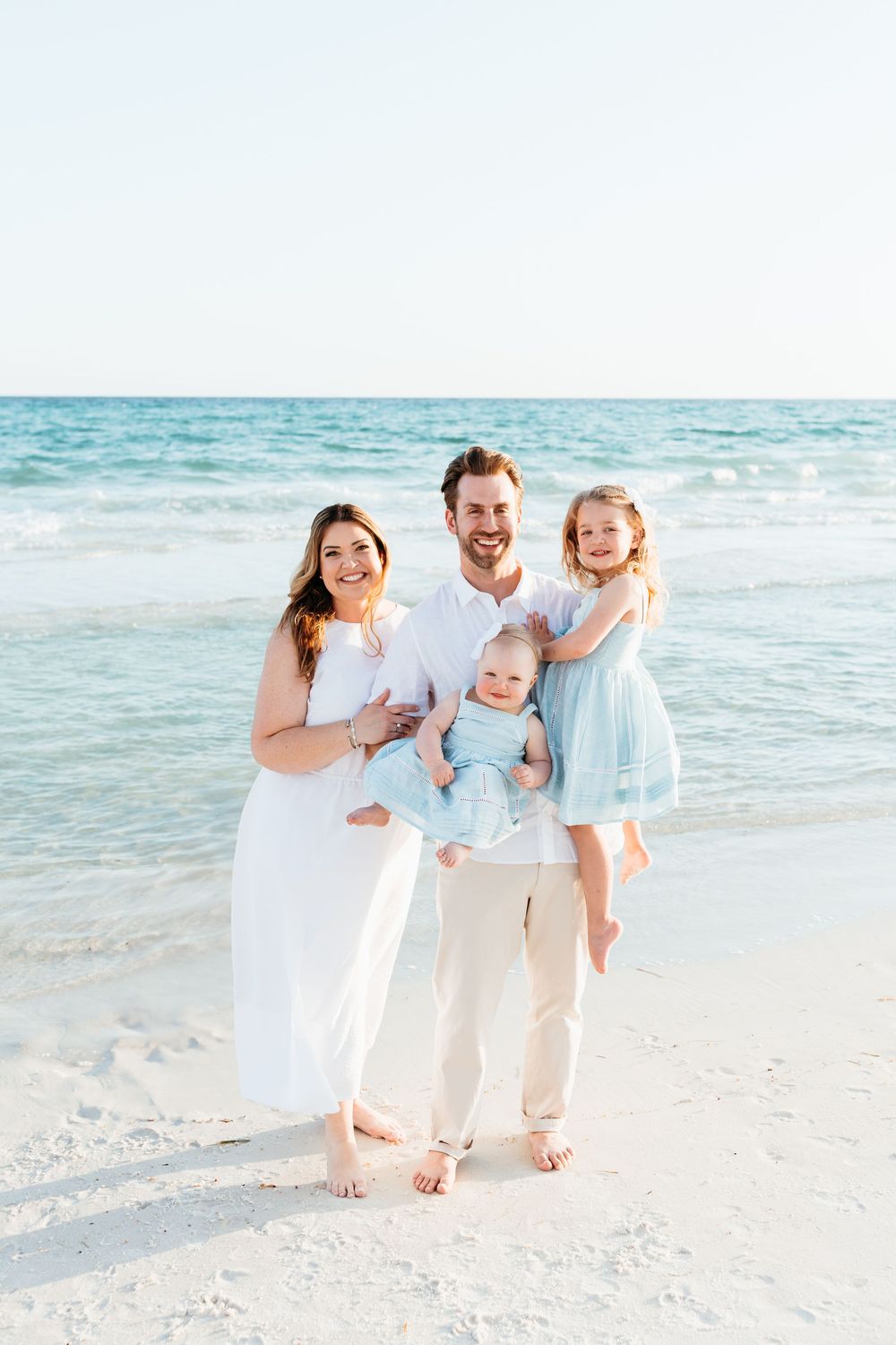 A family in coordinated white and blue outfits poses together on a sunny beach with turquoise ocean waters behind them.