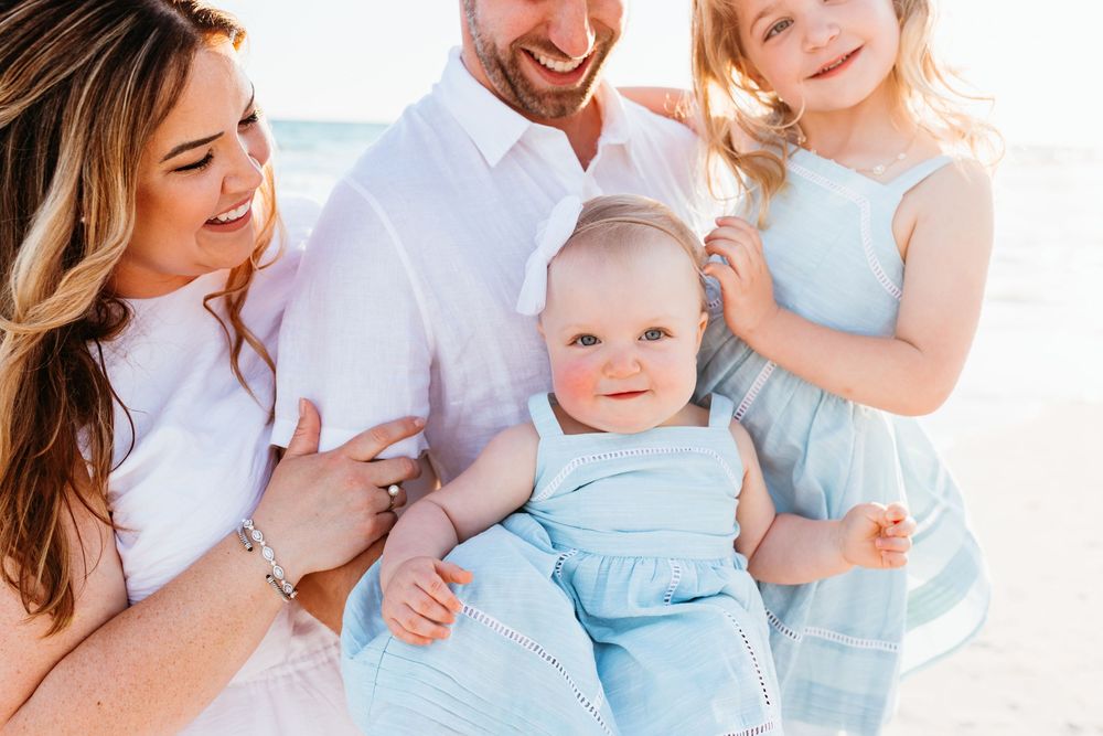 A joyful family group wearing white and light blue outfits poses together outdoors against a bright background.