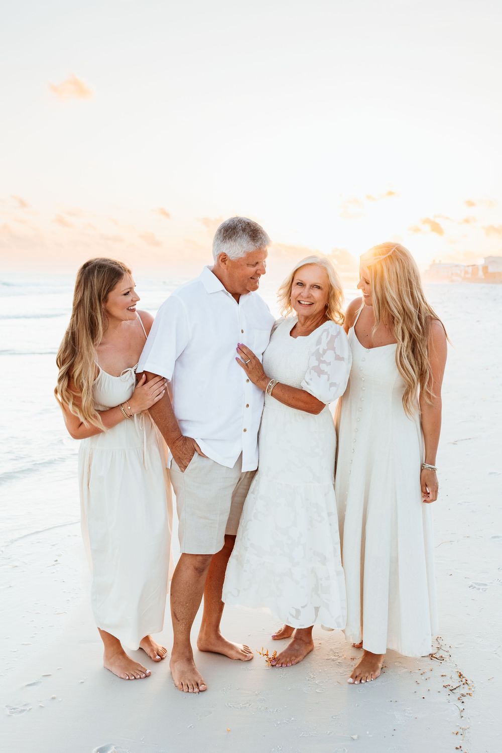 A family of four wearing white outfits smiles together on a beach at sunset.
