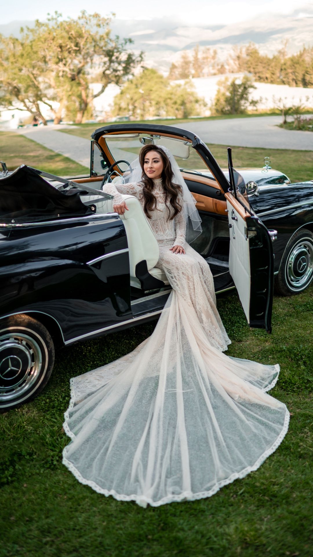 Flowing white wedding dress twirls next to a classic black Mercedes convertible on grass.