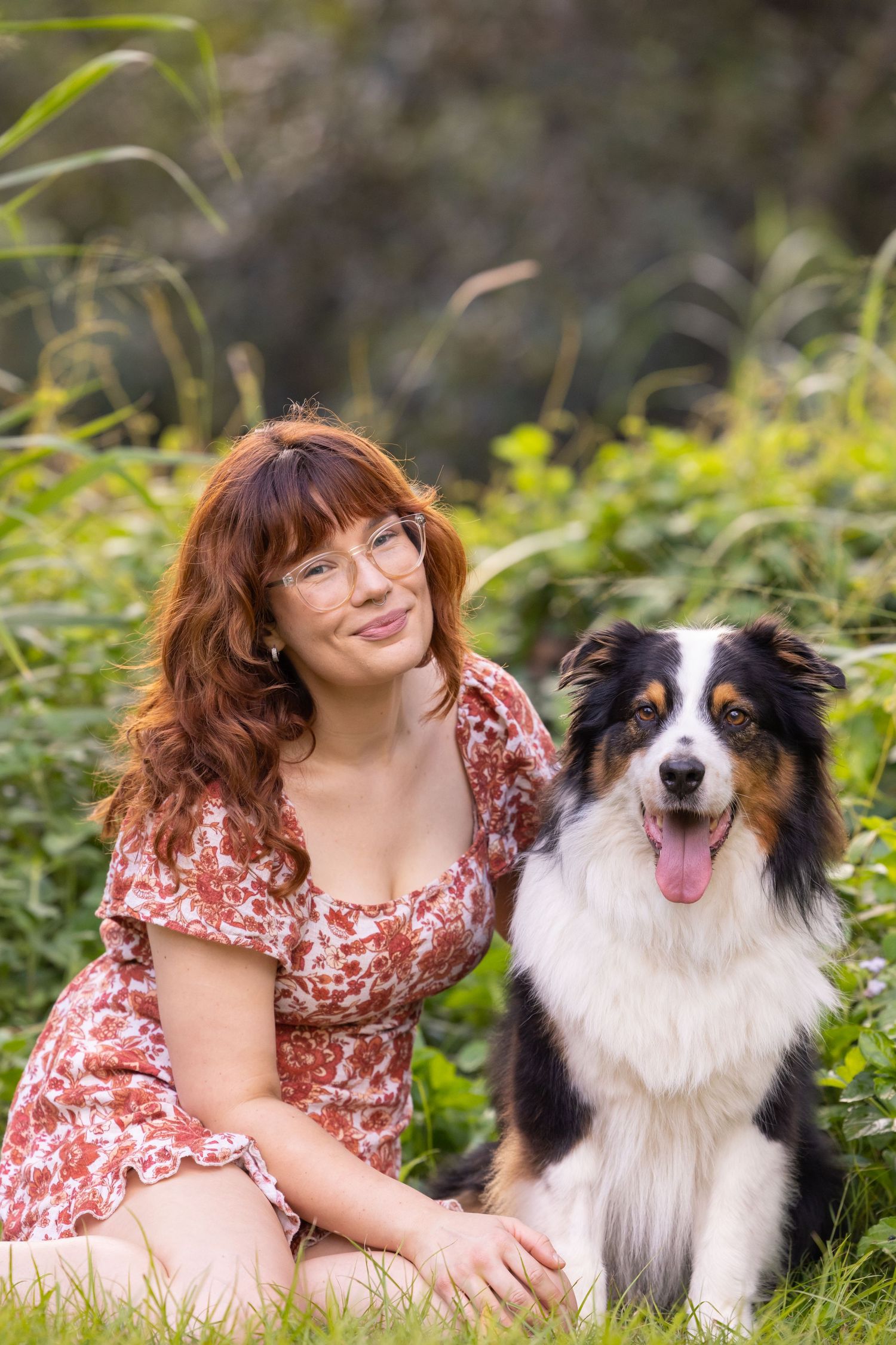 Girl with red wavy hair and front fringe wearing glasses smiling next to her black tri Australian Shepherd dog
