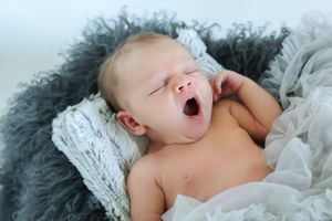 A yawning newborn baby lies on soft white and gray fabric while taking a nap.