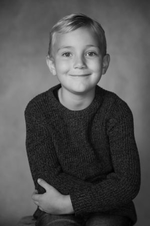Black and white portrait of a young child wearing a dark knit sweater with a gentle smile against a gray background.