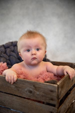 Baby in a rustic wooden crate with pink fabric, photographed against a gray background.
