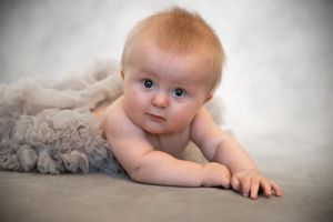 Infant lying on soft gray blanket with bright blue eyes and light hair looking directly at camera.