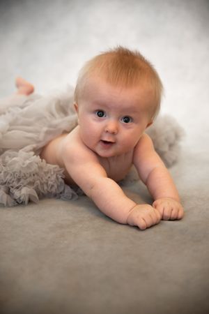 A baby with light reddish hair lies on a soft gray blanket while smiling at the camera.