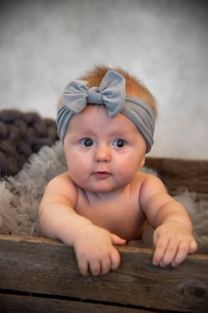 Baby wearing soft gray headband with bow poses laying down on rustic wooden surface with plush blanket.