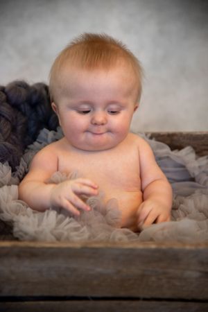 Sweet baby with reddish hair sits in wooden container surrounded by soft grey fabric material.