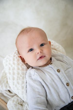 Baby wearing a light colored button-up shirt lies on textured white blanket, gazing upward with bright eyes.