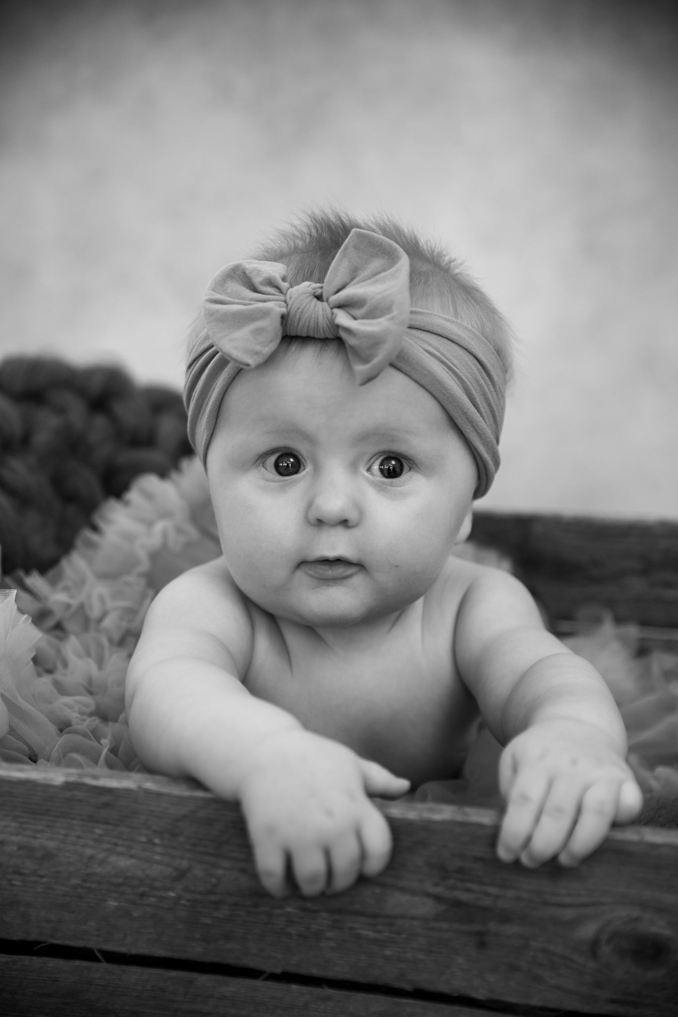 Black and white portrait of baby wearing bow headband while leaning on wooden surface.