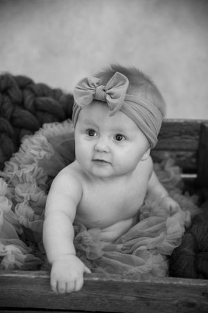 Black and white portrait of a baby wearing a bow headband and tulle skirt while lying on textured furniture.