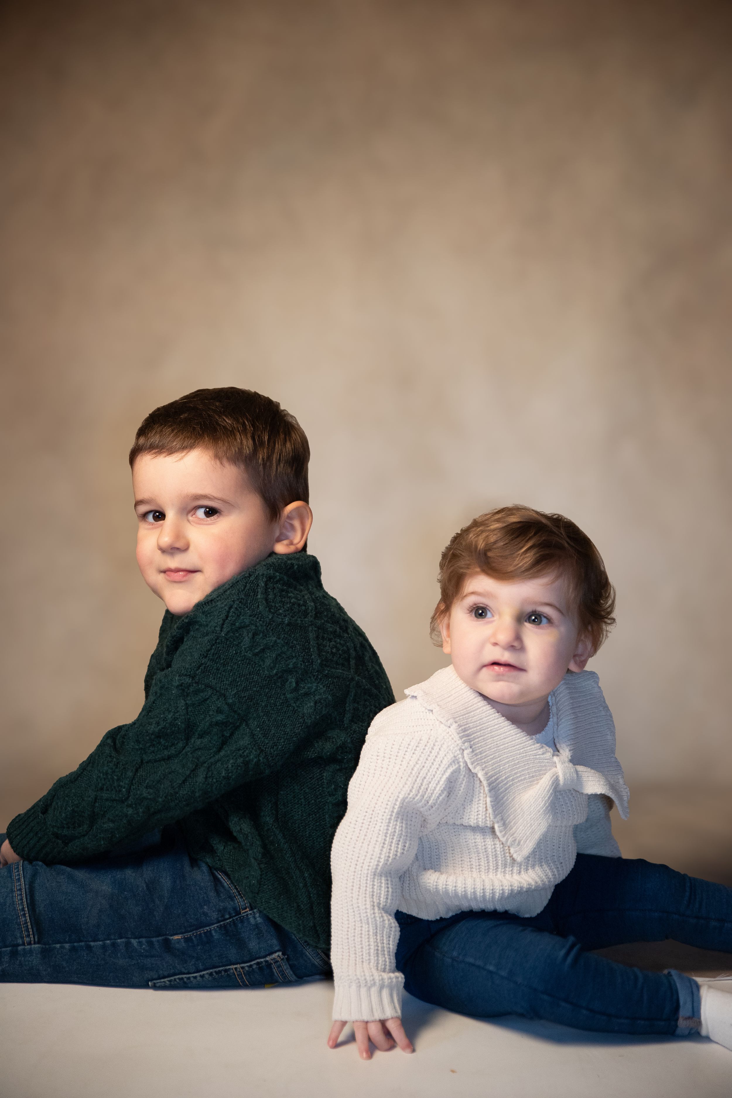 Two children wearing cozy knit sweaters sit back-to-back on white surface against brown studio backdrop.
