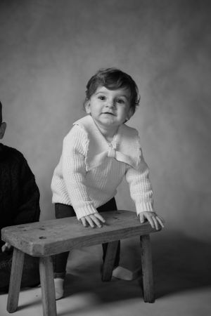 A small child in a white sweater leans over a wooden bench in this vintage black and white studio portrait.