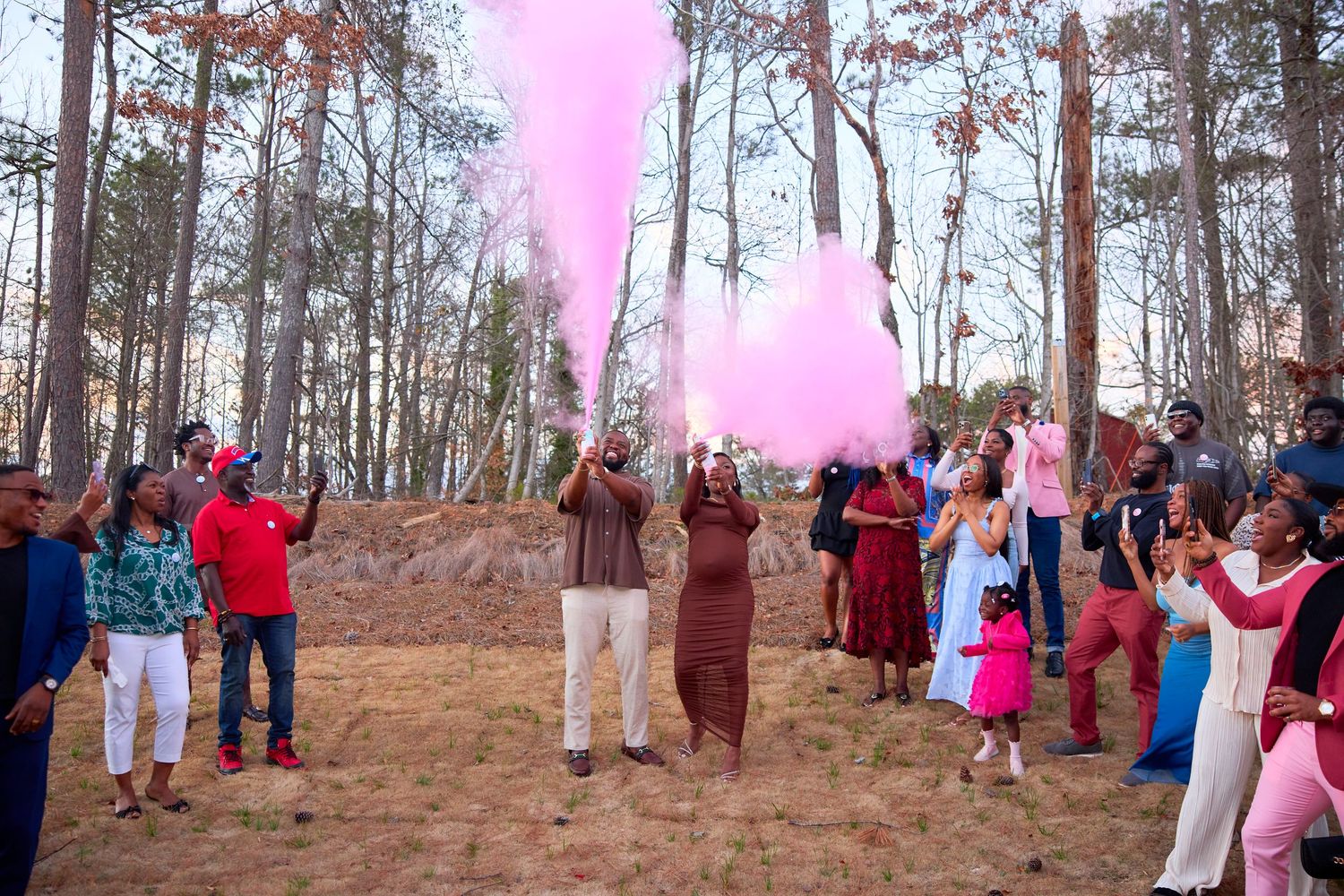 A group of people gather in a wooded area with pink smoke filling the air during an outdoor celebration event.
