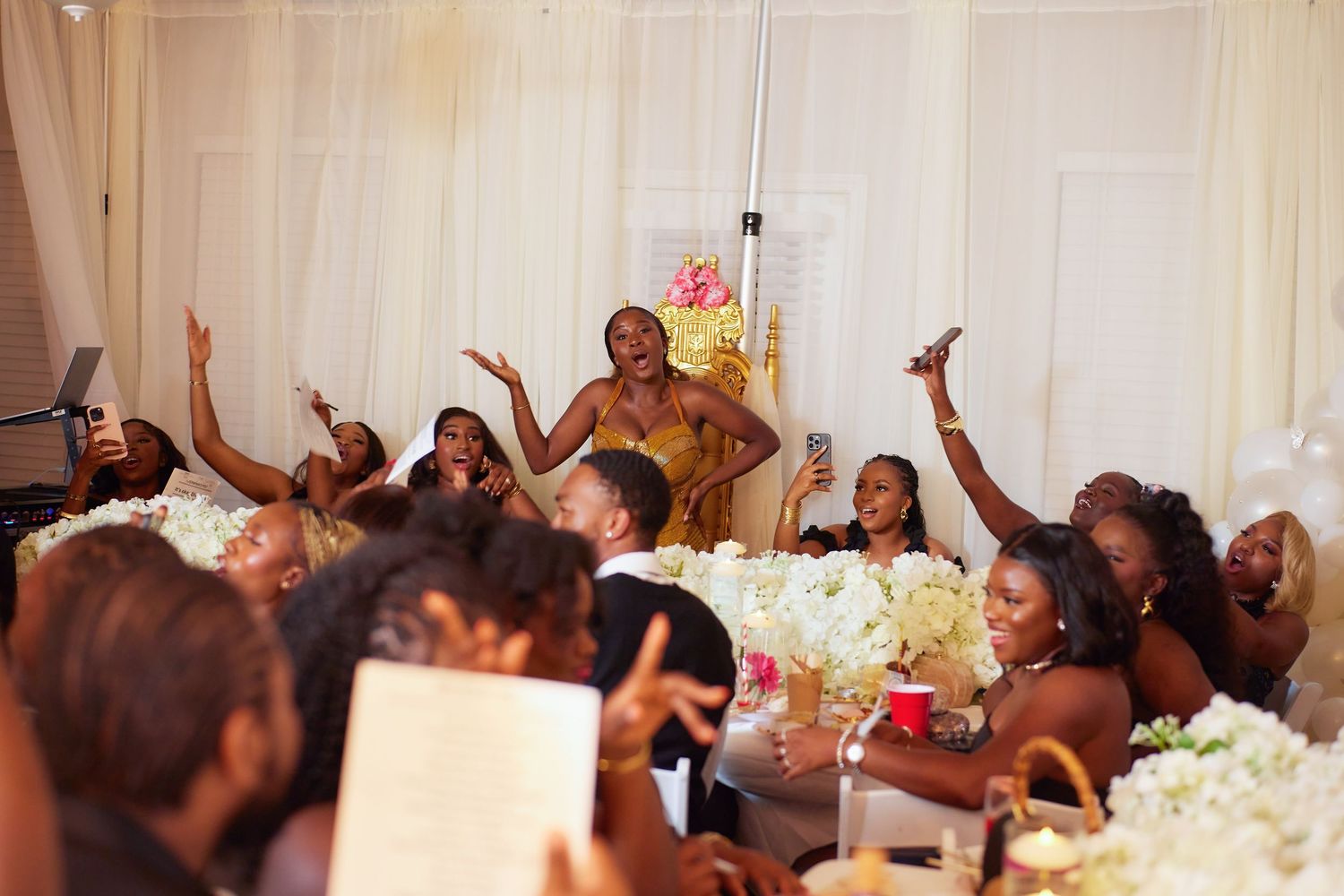 Wedding reception guests raise their hands in celebration at a table decorated with white floral centerpieces.