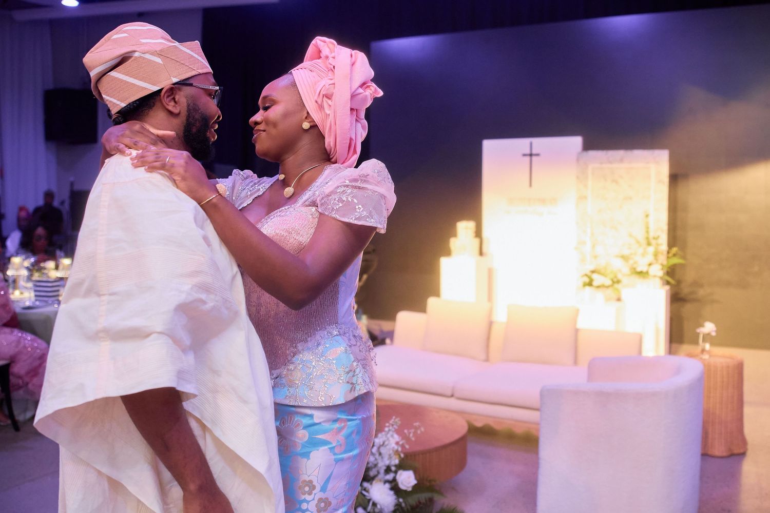 Two people share a joyful moment in traditional white and pink attire during a church ceremony.