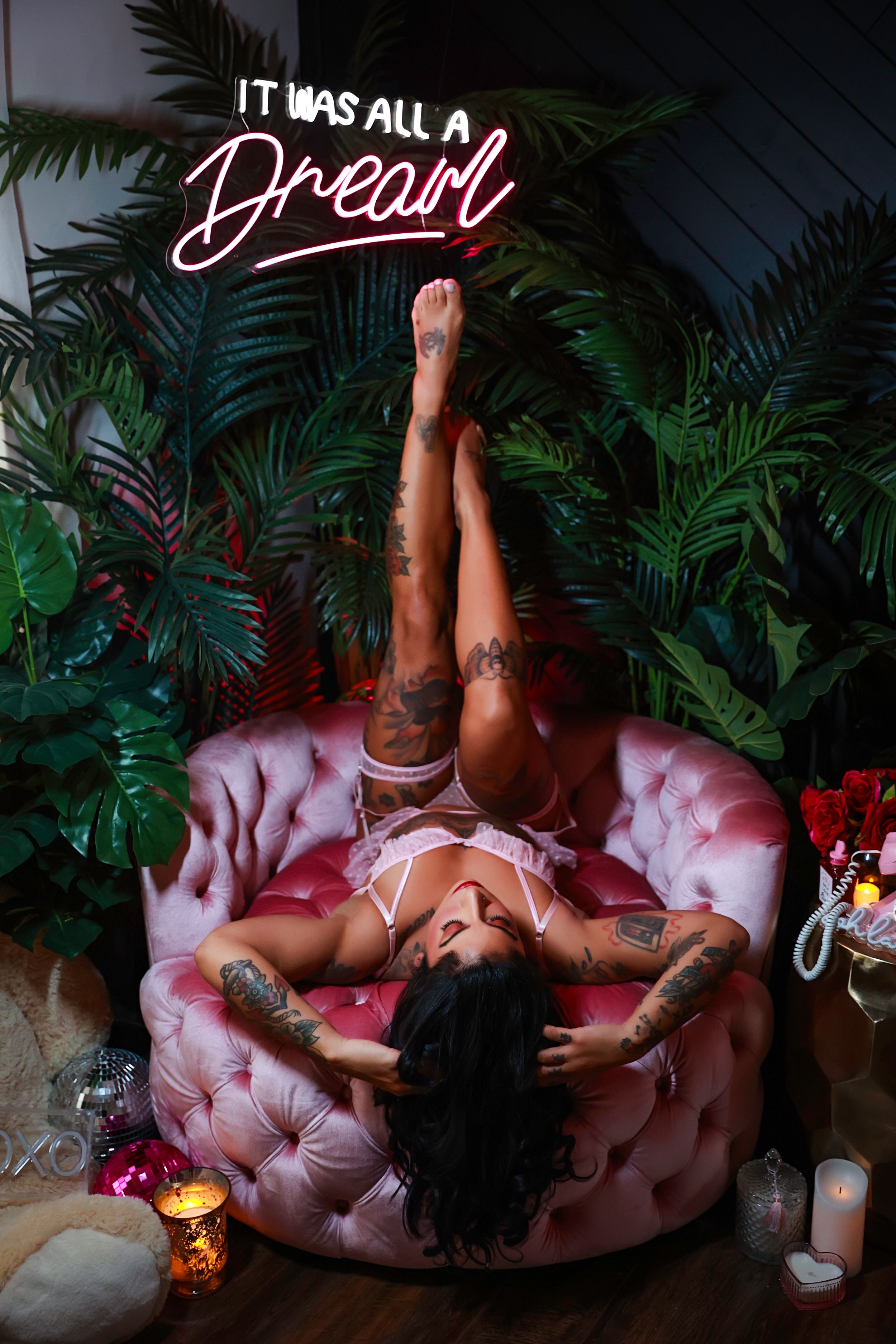 Glamour photo with dramatic pose below a pink neon lighted "it was all a dream" sign and tropical plants against pink tufted couch.