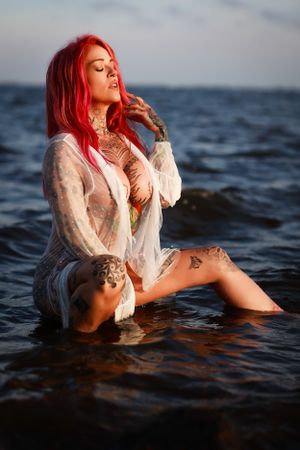 Boudoir Model with bright pink hair poses dramatically in dark ocean water wearing a sheer white top.