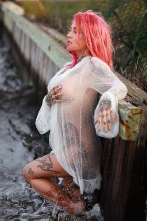 Artistic outdoor boudoir photoshoot featuring sheer white clothing and pink hair against a rustic wooden backdrop standing in the ocean.