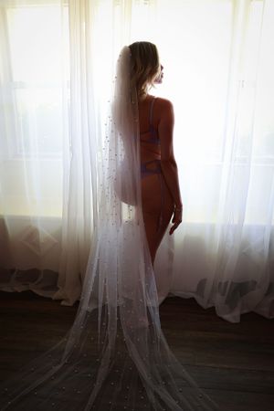 Beautiful woman with long wedding veil and lingerie silhouette figure against sheer white curtains in natural window light.