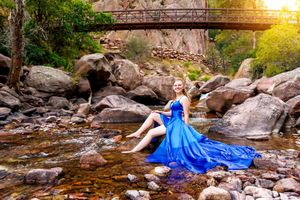 Woman in blue gown sitting on rocks by a stream with a wooden bridge and lush greenery in the background.