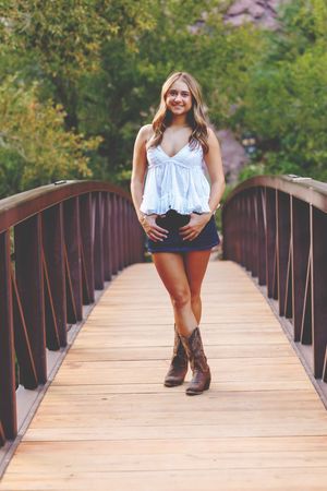 Young woman in white top, denim skirt, and cowboy boots smiling on a wooden bridge surrounded by green trees.