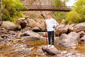 Boy standing on rocks by a mountain stream holding a lacrosse stick, with a wooden bridge and trees in background.