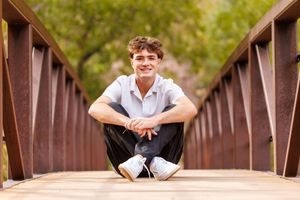 A smiling teenage boy sits cross-legged on a wooden bridge, wearing a white polo shirt and white sneakers.