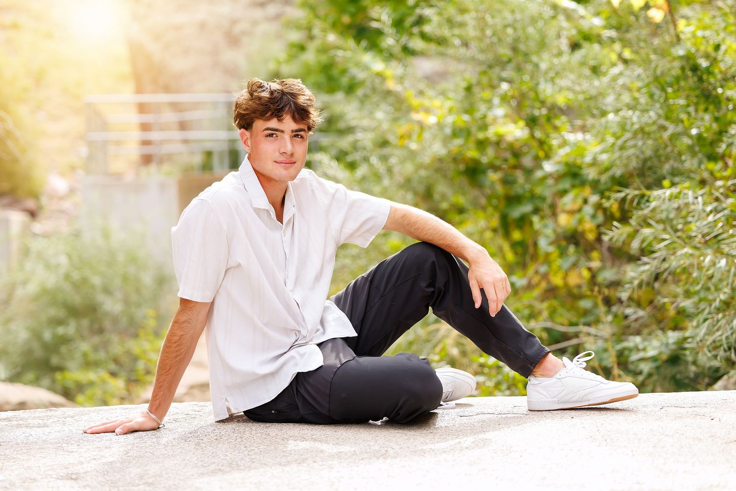 Young man in white shirt and dark jeans sitting on a rock outdoors, surrounded by lush green foliage.
