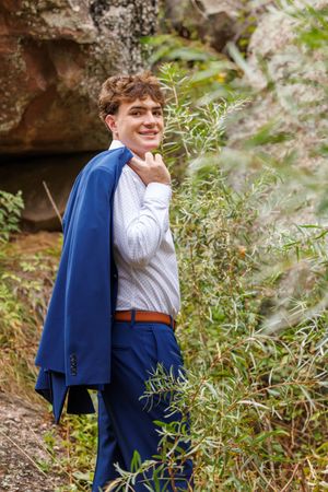 Young man in blue suit holding jacket over shoulder, smiling outdoors near rocks and greenery.